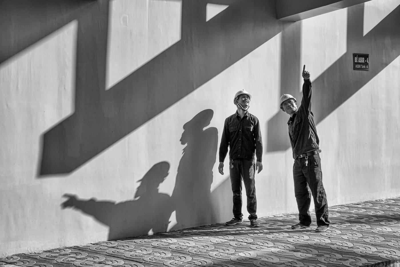 Two workers in hard hats pointing up at a wall striped with shadows